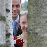 Foto von einem Paar in Hochzeitskleidung mit einem Strauß aus roten und weißen Rosen, der zwischen zwei Baumstämmen steht.
